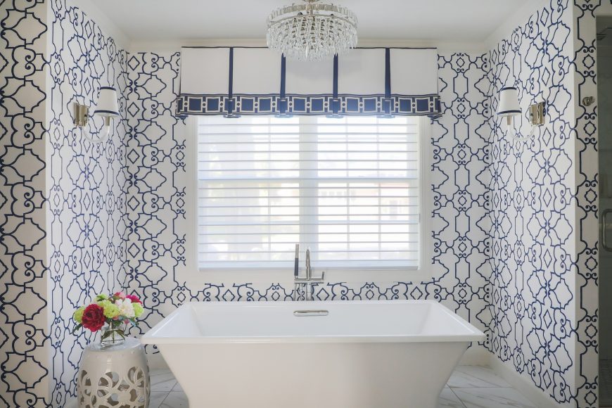 The primary bathroom is a highlight of the renovation. A built-in tub with decking was swapped for an elegant, freestanding soaking tub by Kohler. The navy Ogden wallpaper by Thibaut is a striking accent, along with the patterned valance.