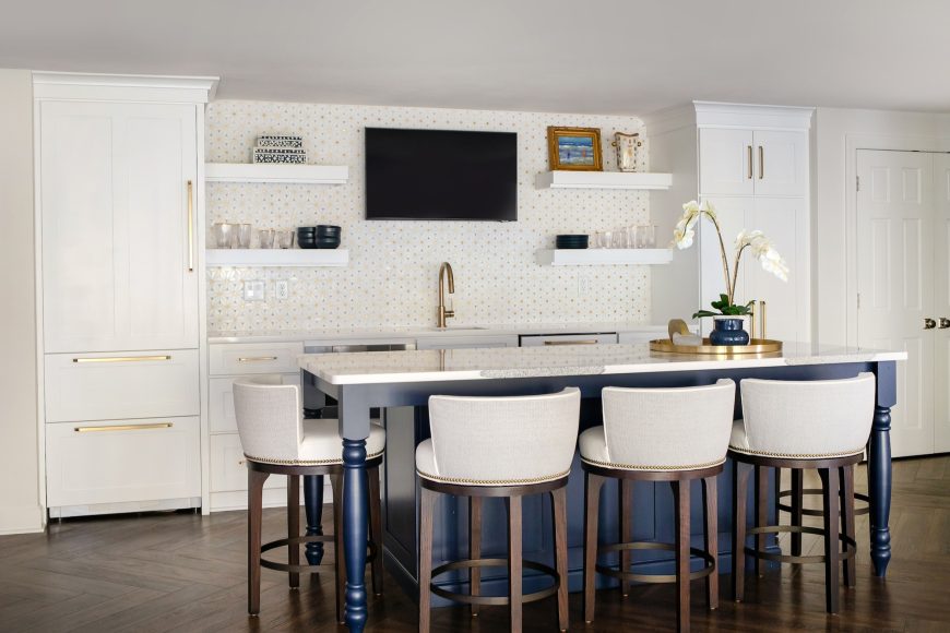 The lower-level kitchenette previously sported dark cabinetry and a soffit. Today, with white cabinetry and sans soffit, it has a much lighter feel. Pops of brass in the faucet, backsplash tile and elsewhere create interest.